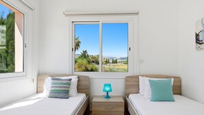 A room with two beds, a window with a view of greenery and hills, and a nightstand with a lamp. - Villa Crystal by Ezoria Villas (Coral Bay)
