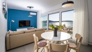 En moderne stue med en beige sofa, et fladskærms-tv, et spisebord med stole og udsigt til et vandareal gennem store vinduer.