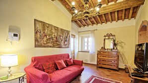 A living room with a red sofa, a wooden dresser, a chandelier, and a large painting on the wall. - Flavia 1 - Vacation Rental with swimming pool in Lucca (Lucca)