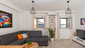 Ein modernes Wohnzimmer mit einem grauen Ecksofa, einem Holzesstisch und einer TV-Konsole. Es gibt zwei Fenster mit Vorhängen, eine Topfpflanze und ein Bild an der Wand.