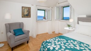 Ein Schlafzimmer mit Bett, einem Sessel, einem Tisch mit Lampe, einem Gemälde und Fenstern mit Blick.