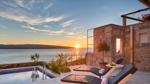 Ein Poolbereich mit Blick auf den Sonnenuntergang über dem Meer, Steinmauern und Liegestühlen.