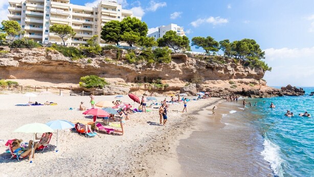 A beach with people sunbathing and swimming, a rocky cliff with trees, and a building in the background. - GALLO Gran villa en playa, piscina y wifi gratis (Miami Playa)