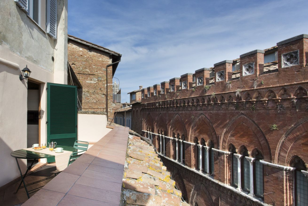 Un stretto vicolo con un muro in mattoni da un lato e un edificio dall'altro. Si nota un tavolo con sedie e una lampada sul lato sinistro.