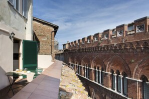 A narrow alleyway with a brick wall on one side and a building on the other. There is a table with chairs and a lamp on the left side.