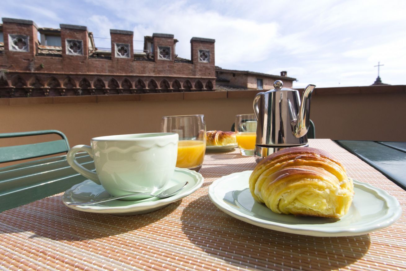 Desayuno con un croissant, café, jugo de naranja y una tetera sobre una mesa con vista a un edificio de ladrillo.