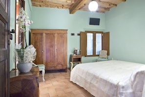 A bedroom with a wooden wardrobe, a bed with a white bedspread, a small table with a vase of flowers, and a window with curtains.