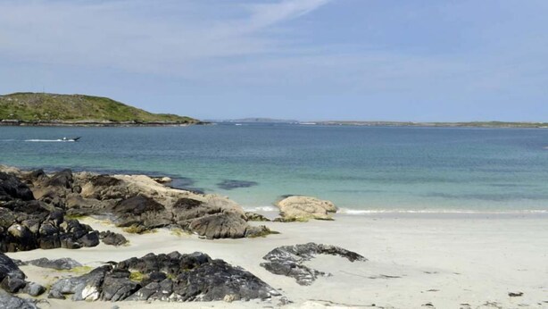 A beach with rocky foreground, calm water, and a distant hill.
