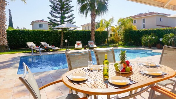 Un espace repas aménagé au bord de la piscine, avec une table dressée pour deux personnes, une piscine et des chaises longues.