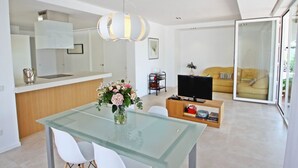 Ein moderner Wohnbereich mit Glastisch, weißen Stühlen, einer Küchenzeile, einer Couch und einem Fernseher.