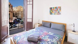 Een slaapkamer met een bed, een nachtkastje, een lamp, een raam met uitzicht op gebouwen en een kleurrijk kunstwerk aan de muur.