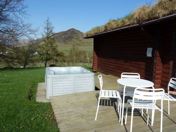 Un jacuzzi, una terraza de madera, sillas de metal blanco y una cabaña con techo de hierba.