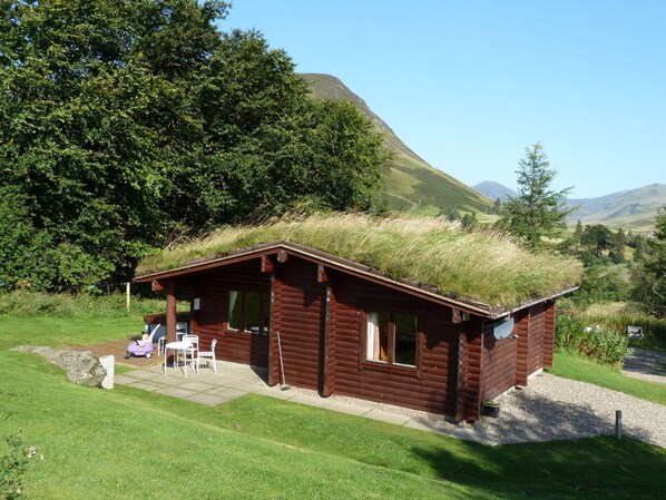 A wooden cabin with a grass roof, surrounded by lush greenery and hills. - Log Cabin with Hot Tub | Sleeps 4 | In the Cairngorm National Park | Accepts Dogs (Spittal of Glenshee)