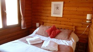 A wooden cabin bedroom with a bed, pillows, a nightstand, a lamp, and a window with curtains.