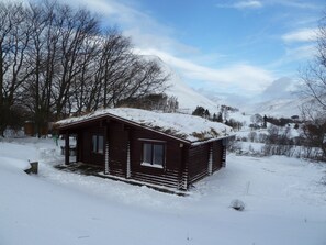 A small wooden cabin with a snow-covered roof, surrounded by a snowy landscape and trees. - Log Cabin with Hot Tub | Sleeps 4 | In the Cairngorm National Park | Accepts Dogs (Spittal of Glenshee)