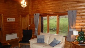A cozy log cabin interior with a beige sofa, a black armchair, a small wooden cabinet, and a Christmas tree.