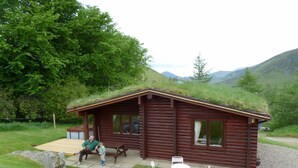A rustic wooden cabin with a grass roof, a porch, and a small outdoor seating area with white chairs.