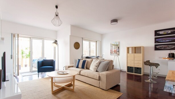A spacious living room with a beige sofa, a round coffee table, and a dining area with a wooden table and bar stools. - JOIVY Stunning apt near Avenida da Liberdade (Lisboa)
