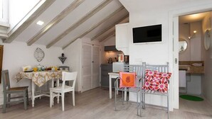 A cozy dining area with a table set for two, a small kitchen area, and a chair with a patterned cushion.