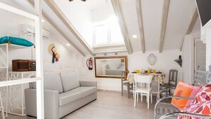 A bright room with a white sofa, a dining table with chairs, a mirror, and a ceiling with wooden beams.