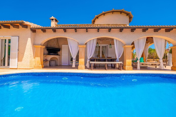 A swimming pool with a covered patio area, a grill, and a table with chairs.