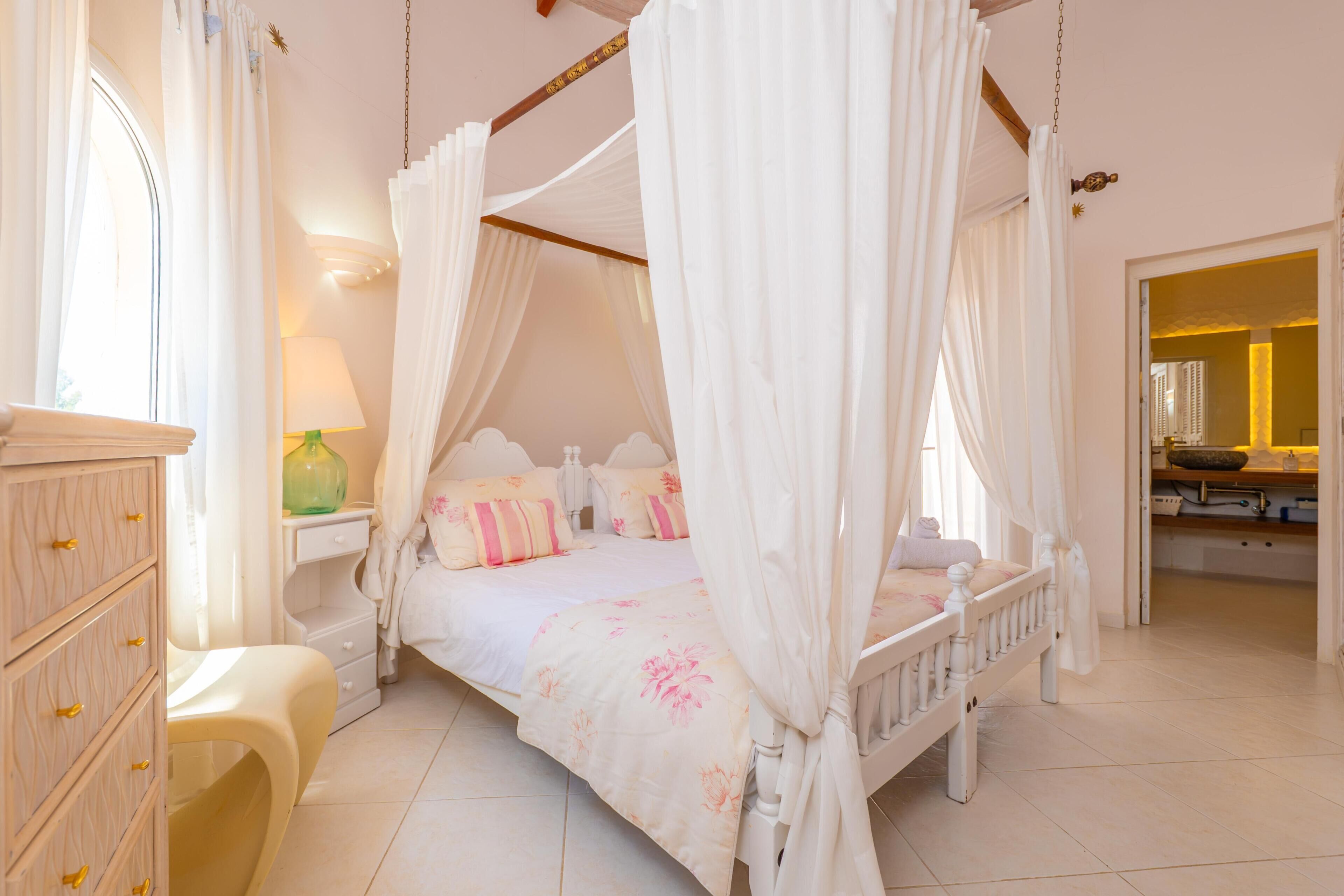Una cama con dosel y cuatro columnas, acompañada de una mesita de noche, un aparador y un espejo en una habitación luminosa.