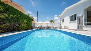 Piscina com espreguiçadeiras e guarda-sóis, cercada por vegetação e um edifício branco.