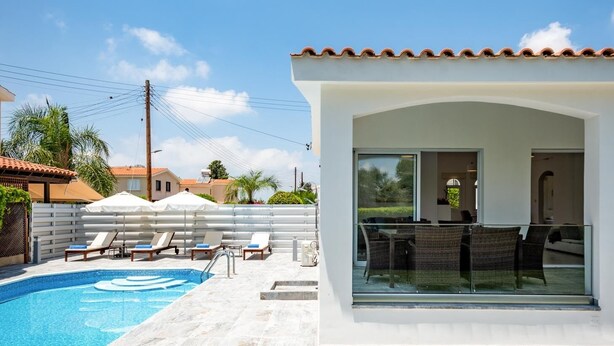 Un área de piscina moderna con un cielo azul despejado, piscina, sillas de descanso y un porche de vidrio con mobiliario exterior.