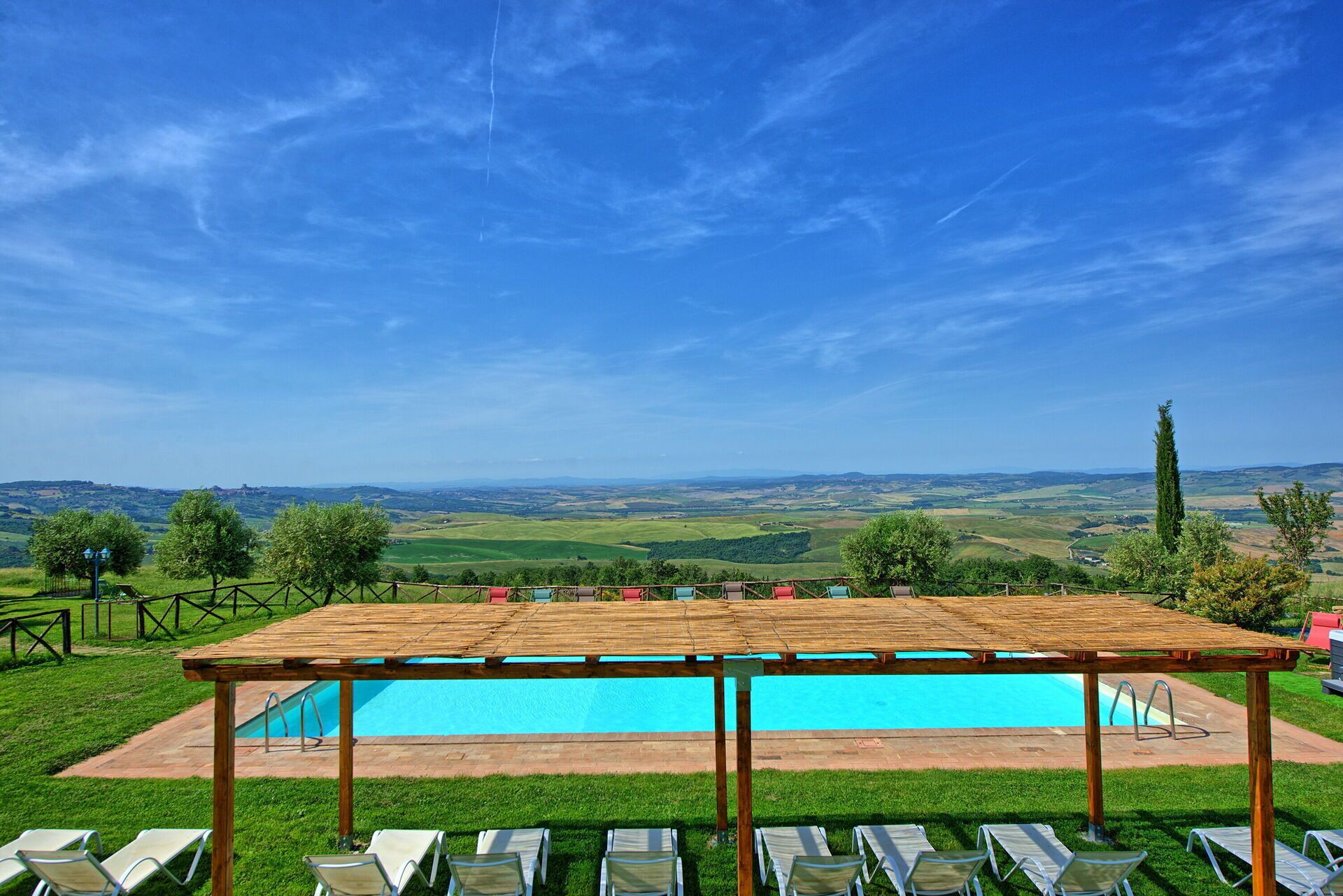 A pool area with a wooden pergola, lounge chairs, and a view of rolling hills.