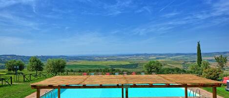 A pool area with a wooden pergola, lounge chairs, and a view of rolling hills.