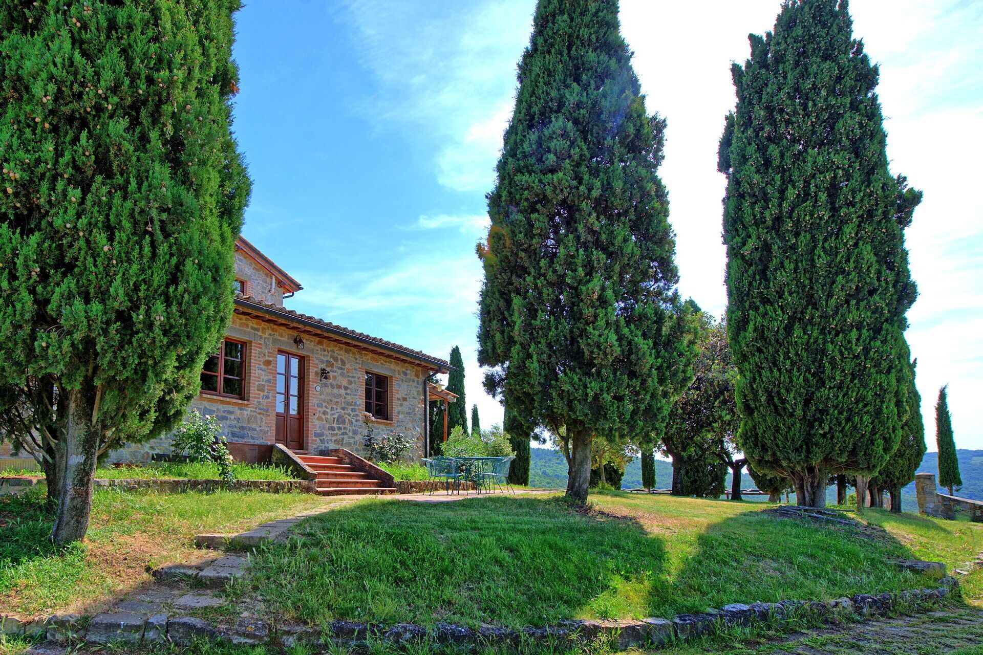 A stone house with a red door, surrounded by well-maintained grass and tall cypress trees.