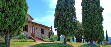 A stone house with a red door, surrounded by well-maintained grass and tall cypress trees.