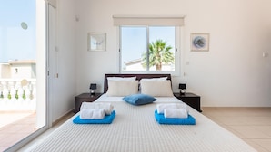 A bedroom with a large bed, two bedside tables, a window with a view of palm trees, and a balcony. - Villa Alysia by Ezoria Villas (Paphos)