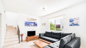 A modern living room with a grey sofa, a flat-screen TV, and a wooden coffee table. A staircase is visible on the left, and there are two windows with artwork on the walls. - Villa Alysia by Ezoria Villas (Paphos)