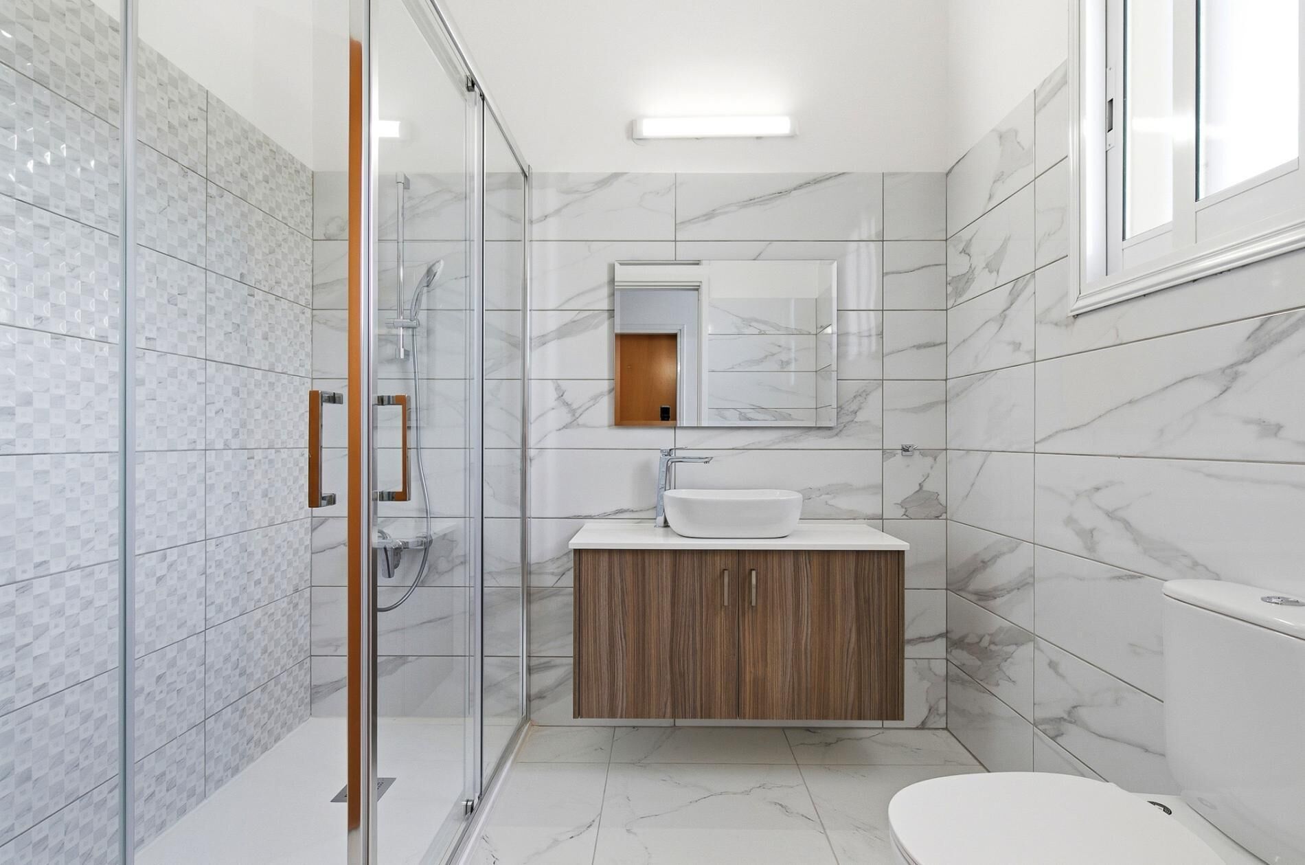 A modern bathroom with a glass shower enclosure, a wooden vanity, a marble-tiled floor, and a toilet.