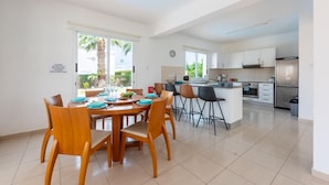 A modern kitchen with white cabinets, a stainless steel refrigerator, and a dining area with wooden chairs and a table set for four. - Villa Alysia by Ezoria Villas (Paphos)