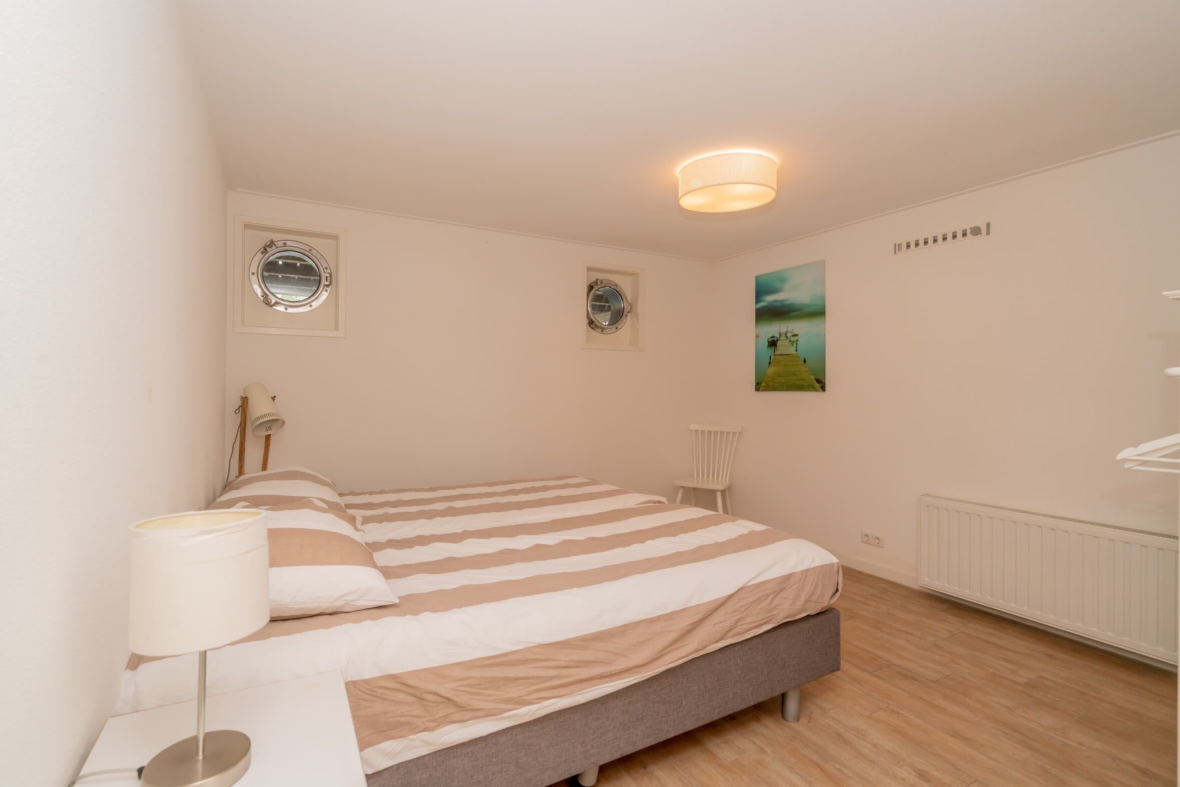 Een slaapkamer met een bed, een bedlampje, twee ronde ramen, een radiator en een schilderij aan de muur.