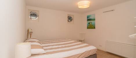 Een slaapkamer met een bed, een bedlampje, twee ronde ramen, een radiator en een schilderij aan de muur.