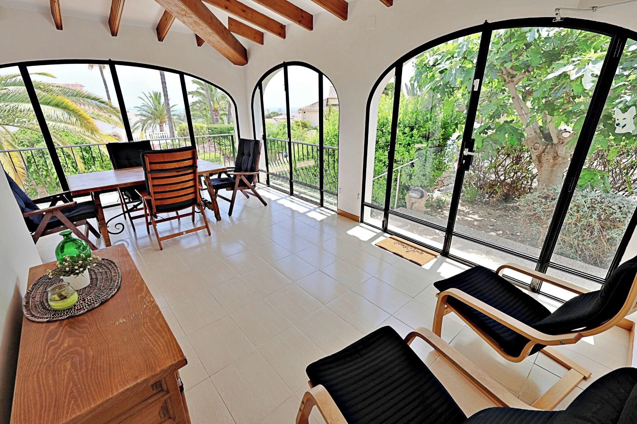 A room with a dining table, chairs, a sofa, and large arched windows offering a view of greenery and buildings.