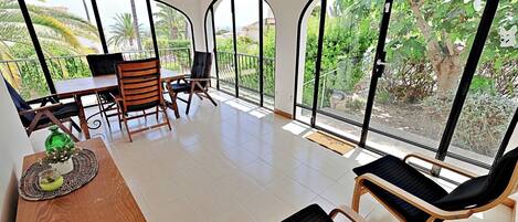 A room with a dining table, chairs, a sofa, and large arched windows offering a view of greenery and buildings.