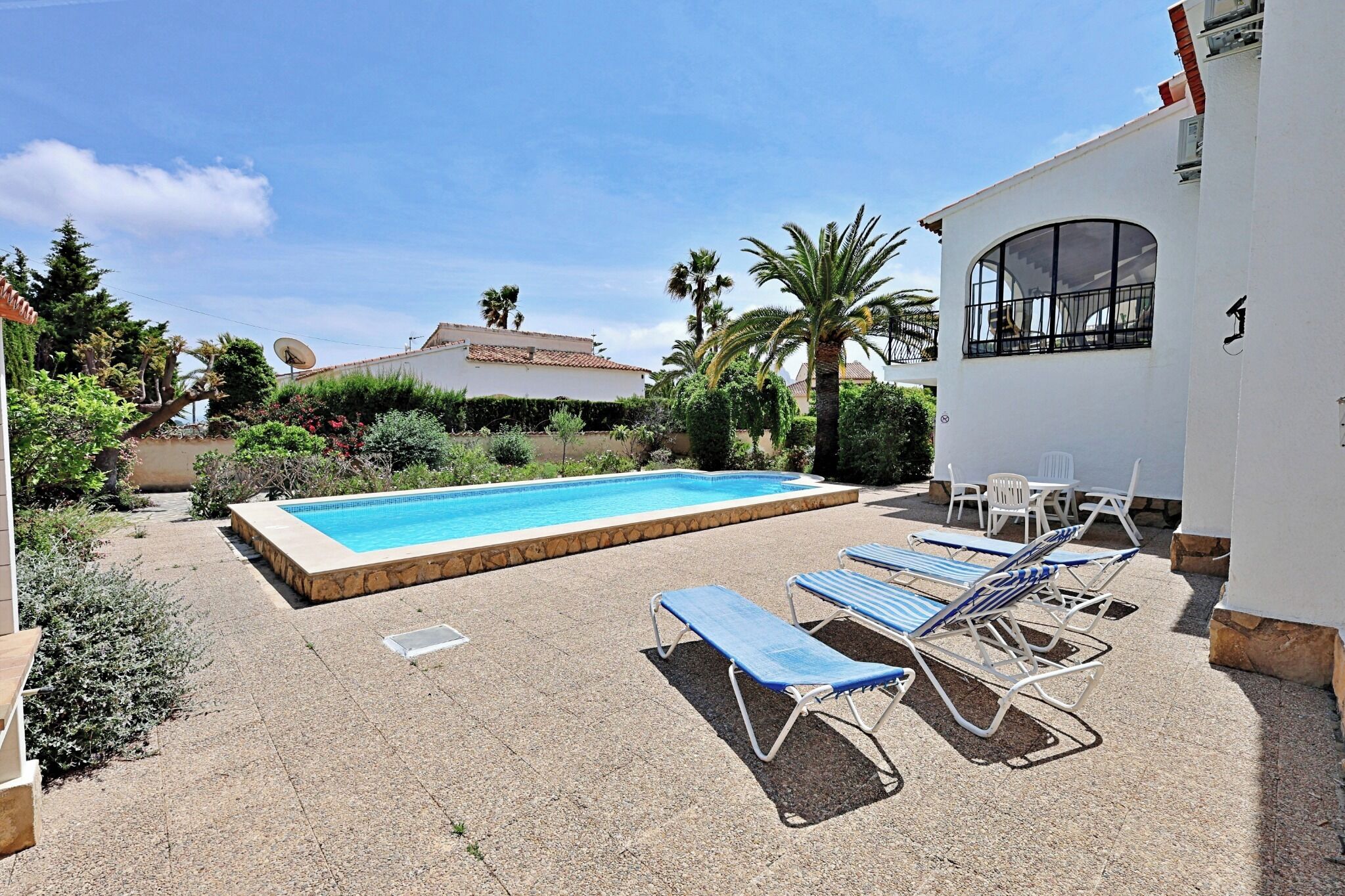 Une piscine entourée de chaises longues, un bâtiment blanc doté d’une fenêtre, et une végétation luxuriante.