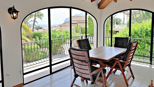 Een balkon met een houten tafel en stoelen, grote gewelfde ramen en uitzicht op groen en gebouwen.