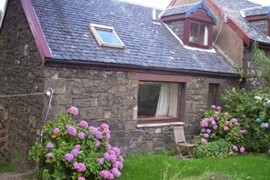 A stone house with a sloped roof, a small window with a curtain, and a wooden chair in the garden. - Steading Holidays - The Byre (Acharacle)