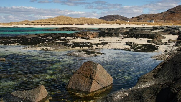 Une plage rocheuse avec une eau claire, des zones de sable et des collines qui se dessinent à l’horizon.