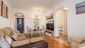 A living room with a sofa, a TV, a dining area with chairs, and a kitchen in the background. - Las Dunas - Punta del Moral Beach PLHD145P118 (Punta del Moral)