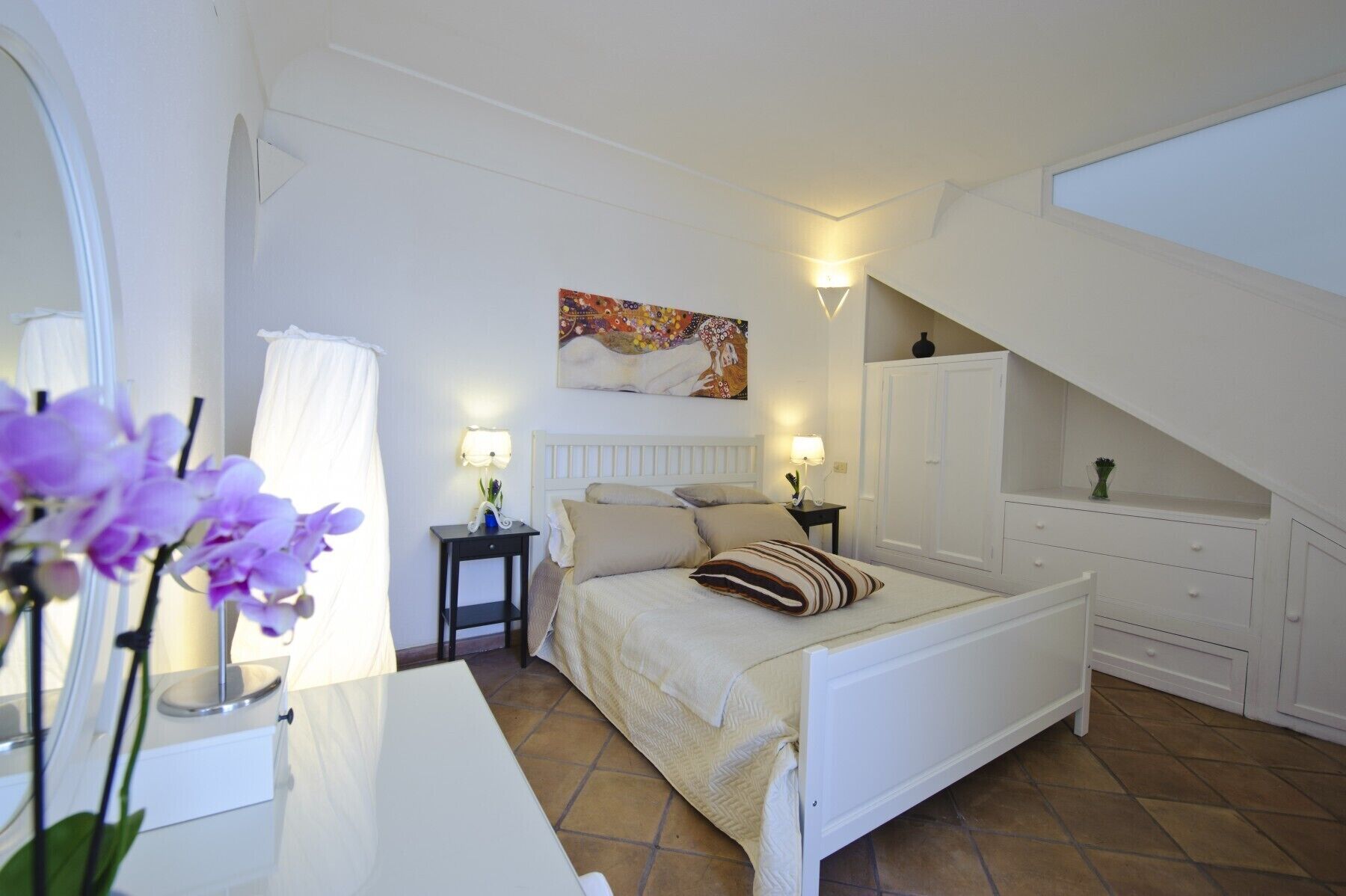 A bedroom with a bed, bedside tables, a painting, and a staircase.