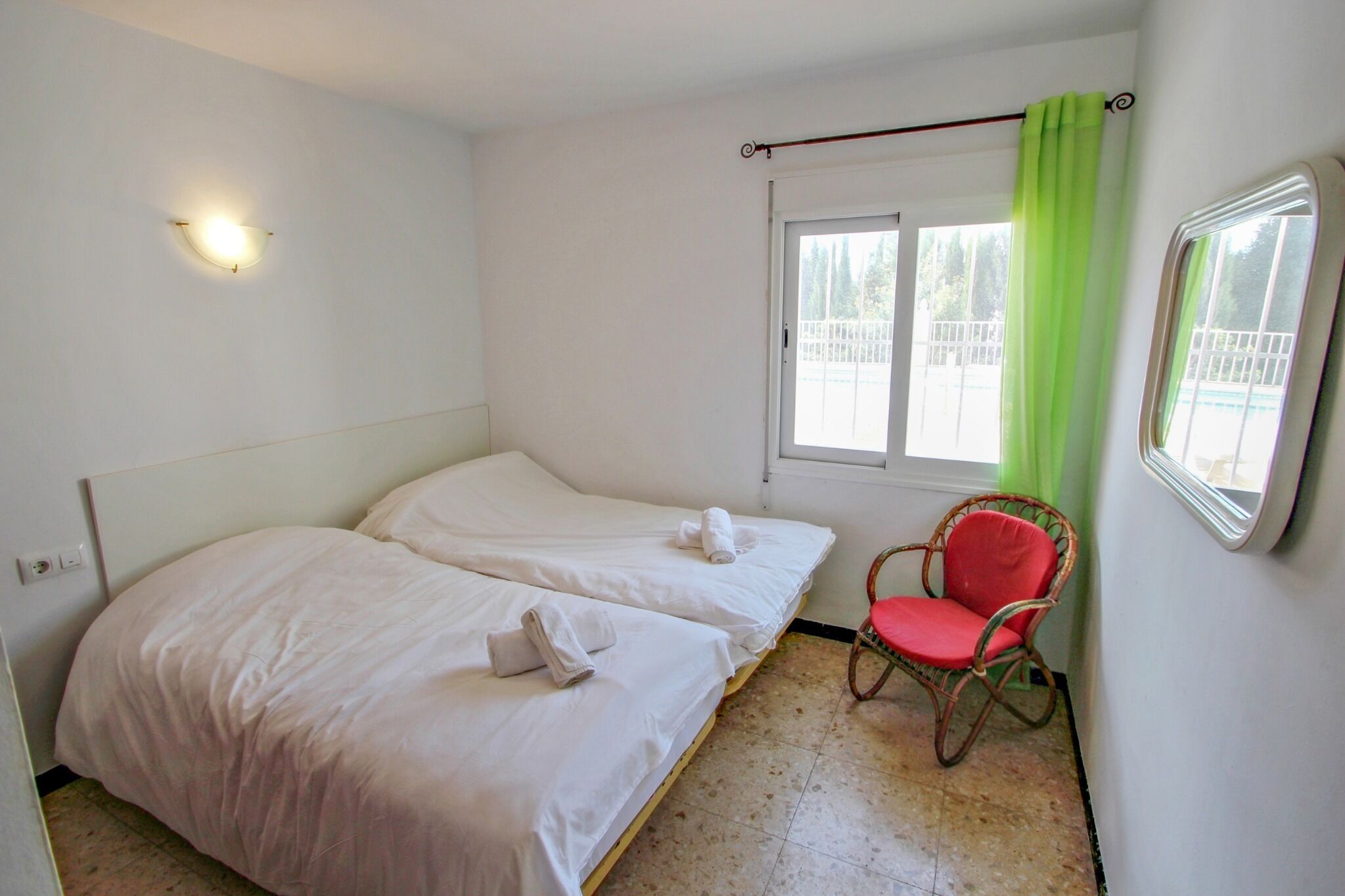A room with two single beds, a red chair, a window with a green curtain, and a wall-mounted light fixture.