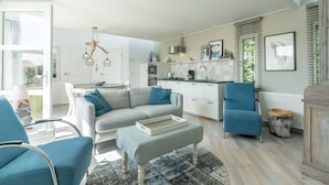 A modern kitchen with white cabinets, a dining area with a grey sofa and blue chairs, and a living room with a patterned rug.