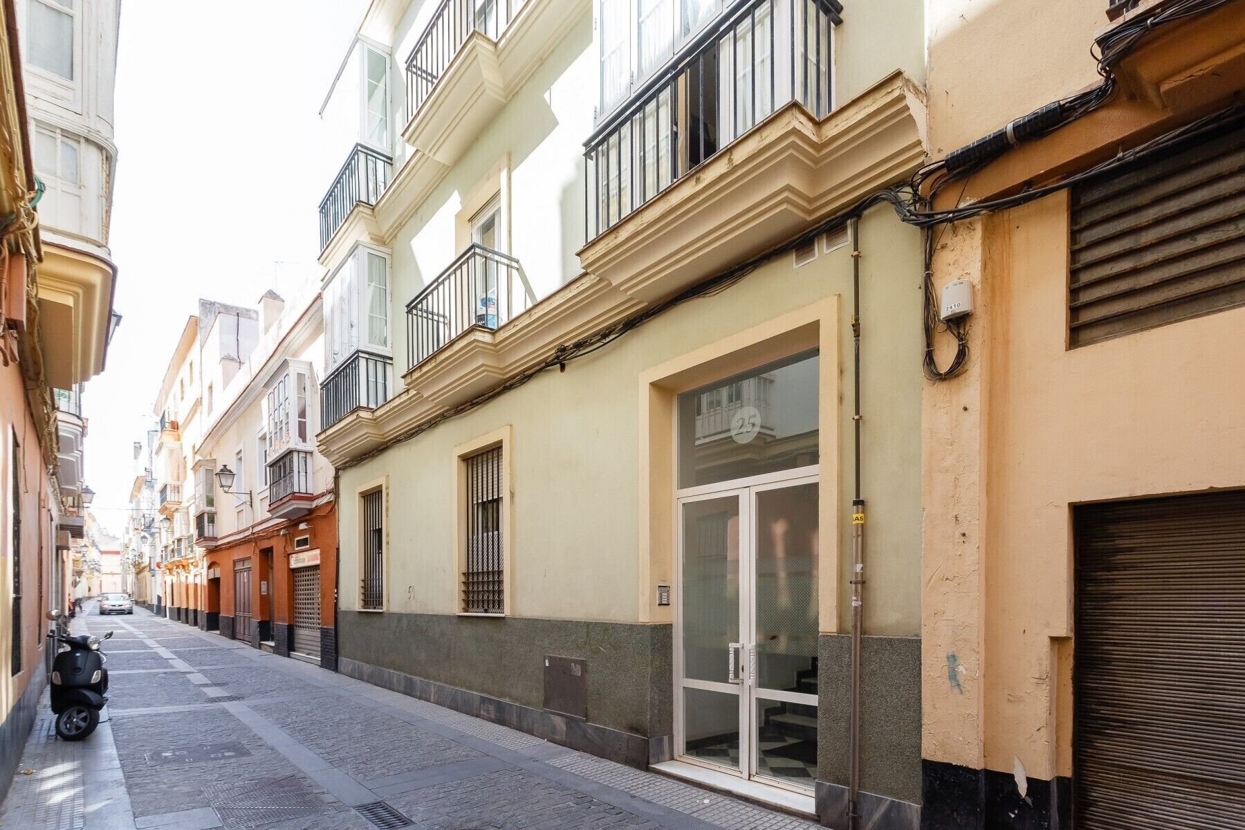 A narrow street lined with multi-story buildings, featuring balconies, large windows, and a closed shop with a glass door.