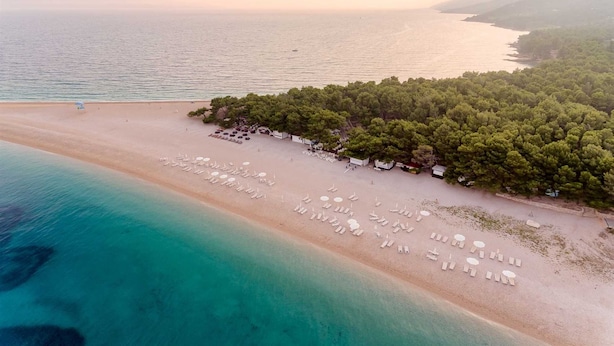Vista aèria d'una platja amb fileres de gandules i para-sols, un grup dens d'arbres i una mar tranquil·la.